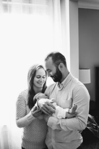 mom and dad smiling at newborn baby next to window