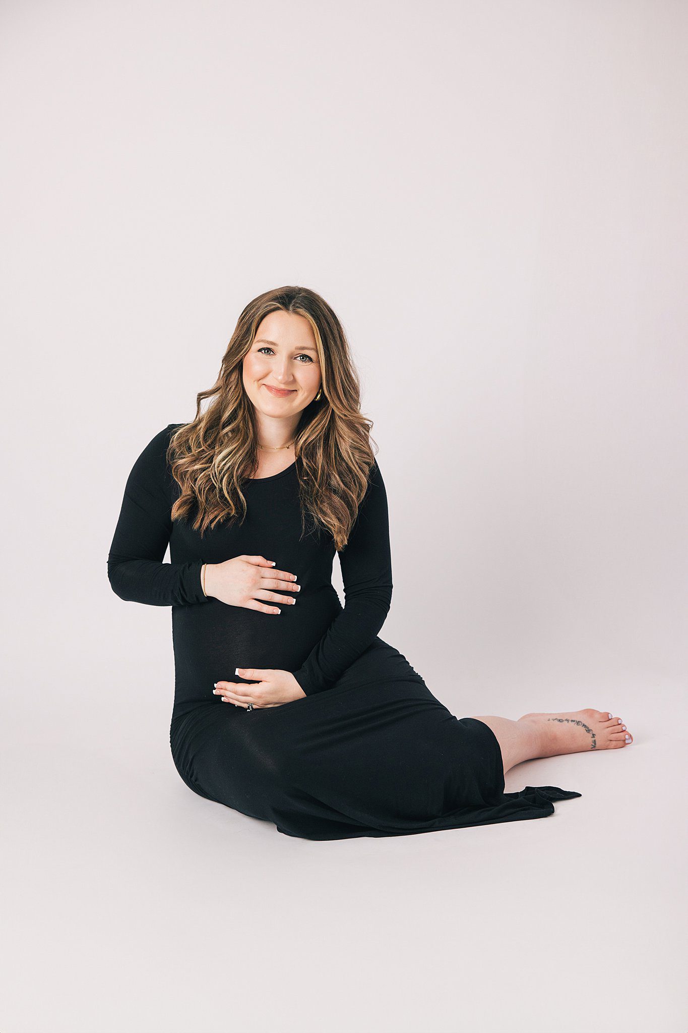 A pregnant woman sits across a studio floor holding her bump and smiling in a black maternity gown
