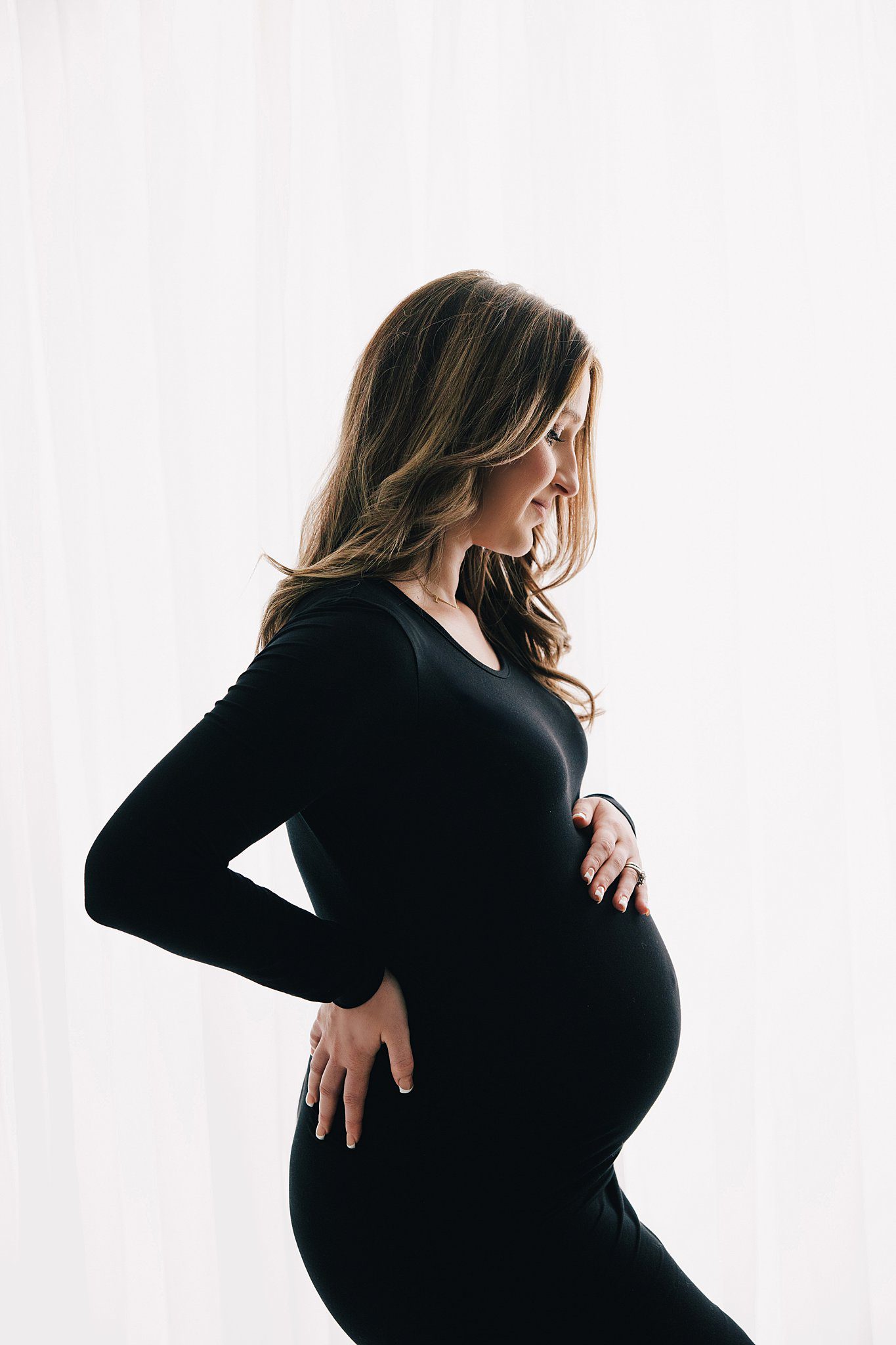 A mom to be in a black maternity gown stands in a window smiling down at her bump after visiting baby shower venues raleigh nc