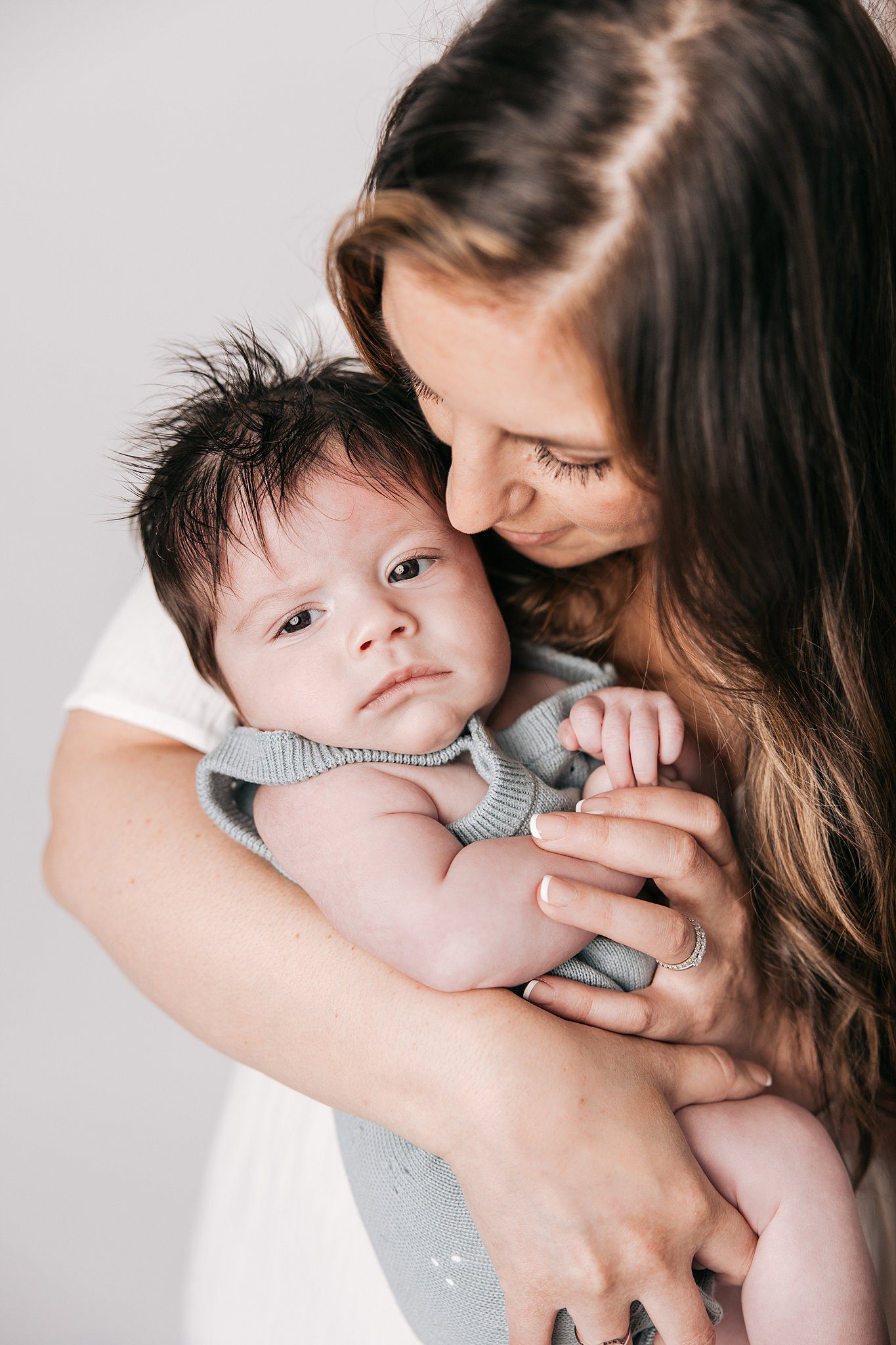 A mother snuggles her infant child while standing in a studio in a white dress