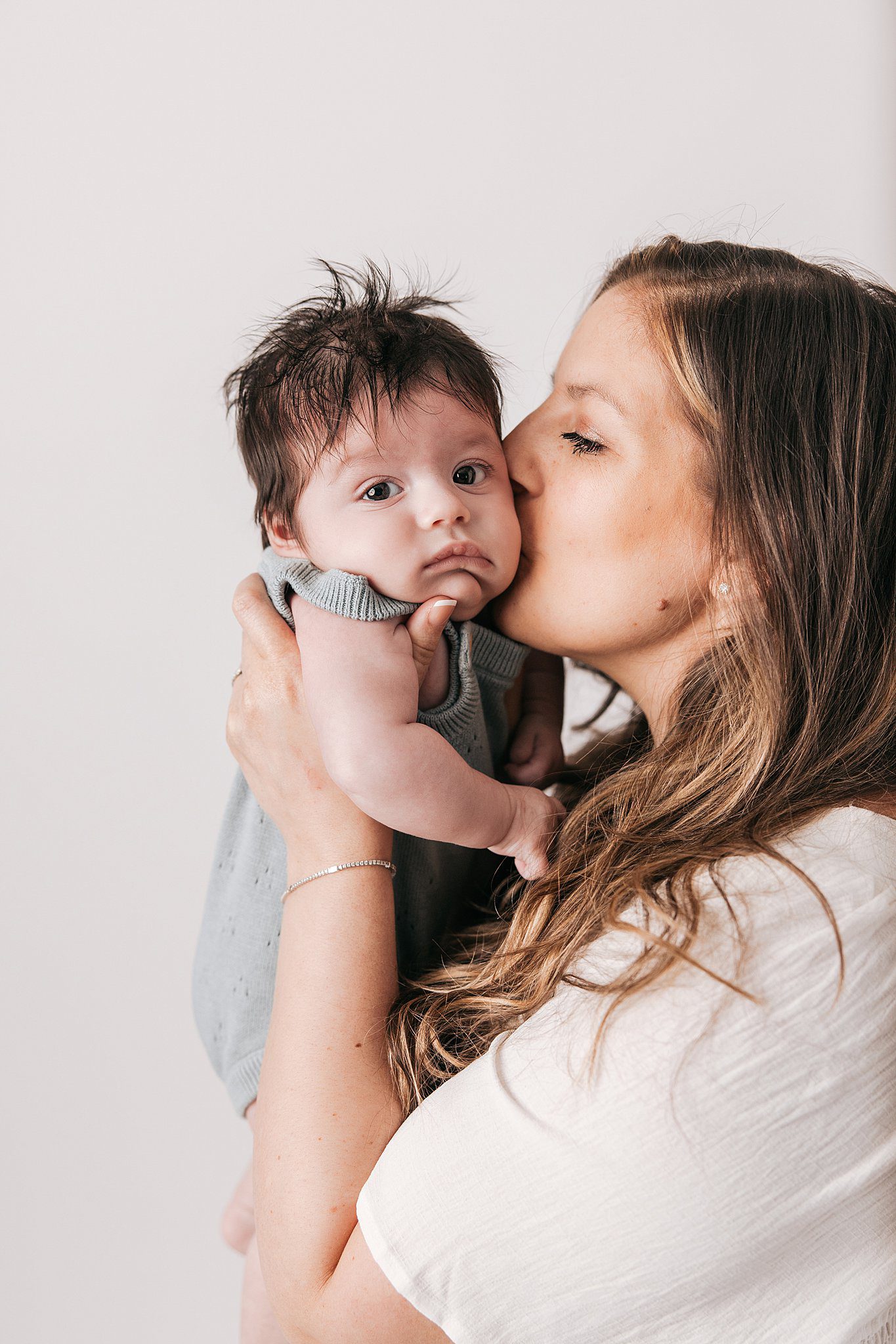 A happy new mother kisses her newborn baby while standing in a studio in a white dress after some mommy and me classes raleigh nc