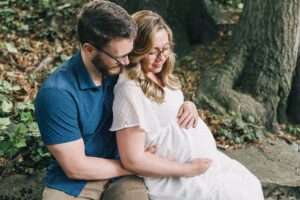 Happy expecting parents sit on a stone wall snuggling and holding the bump