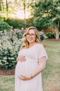A mom to be in a white maternity dress stands in a rose garden holding her bump at sunset after a prenatal massage raleigh