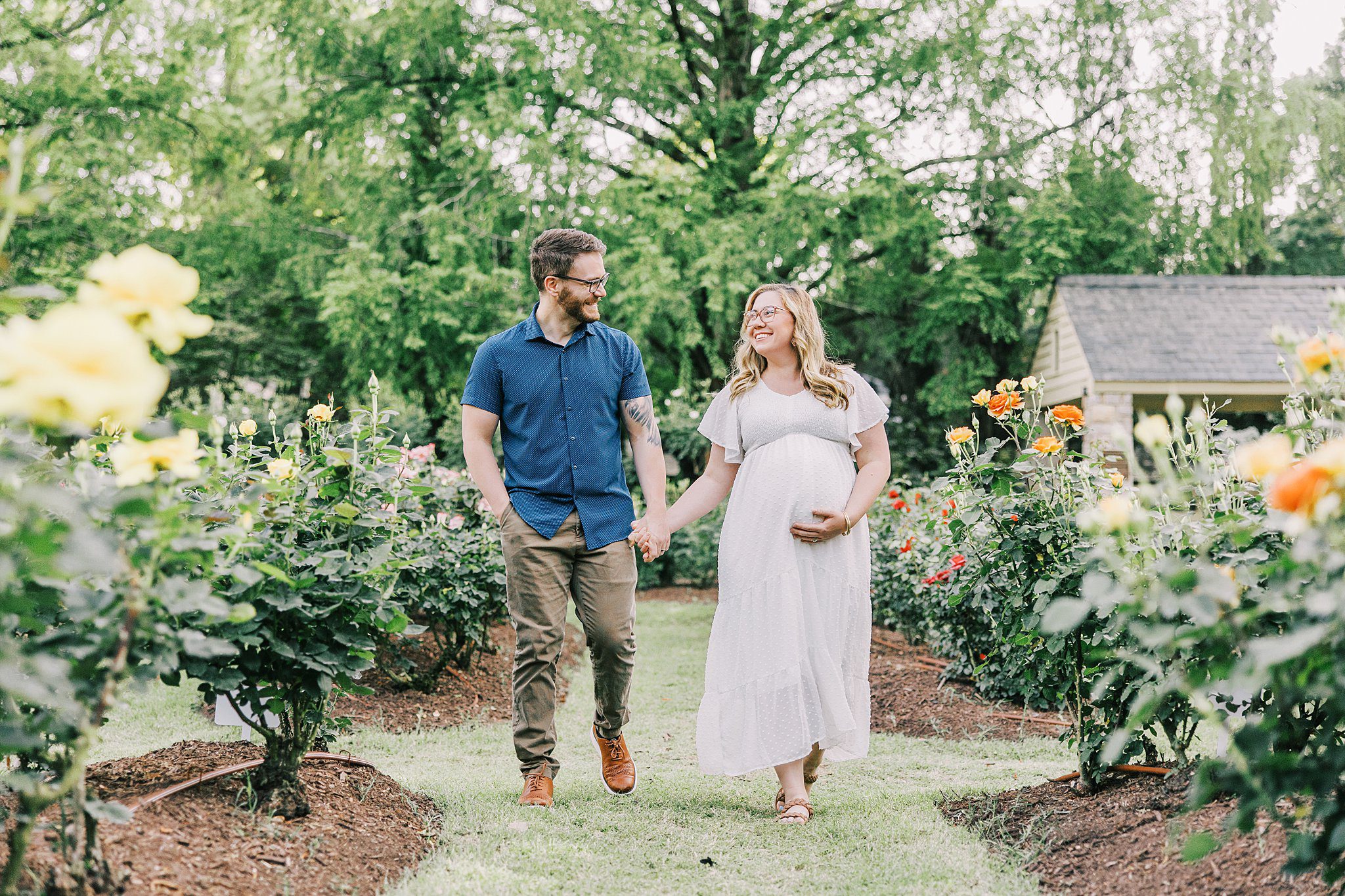 A happy expecting couple holds hands while walking through a colorful rose garden after a prenatal massage raleigh