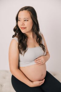 A happy mother to be sits on a bench in a studio holding her bump with a white shirt pulled up