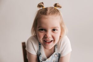 A toddler girl smiles big while sitting on a wooden chair in a studio in denim overalls and pigtails
