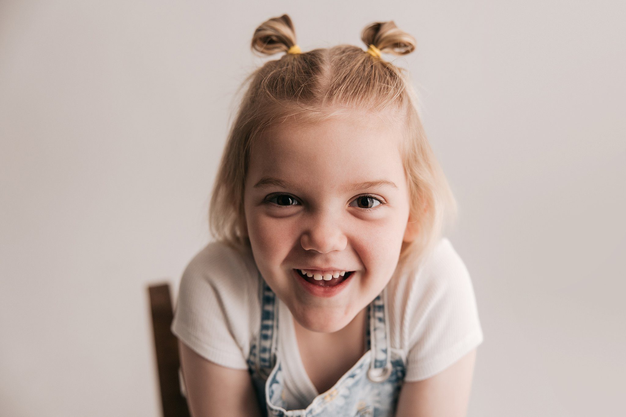 A toddler girl smiles big while sitting on a wooden chair in a studio in denim overalls and pigtails