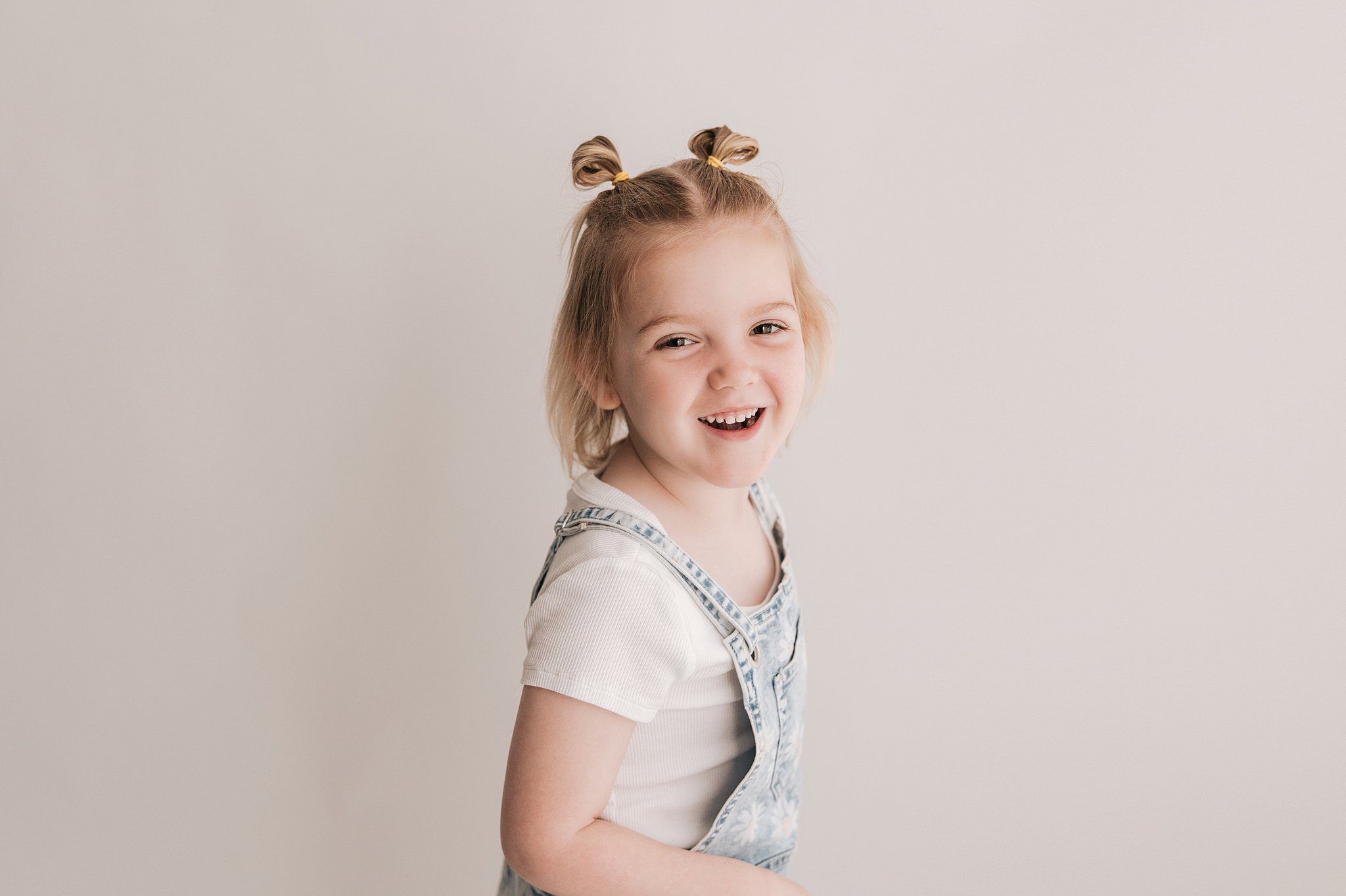 A toddler girl with short pigtails and denim overalls laughs while playing in a studio after meeting with raleigh pediatricians