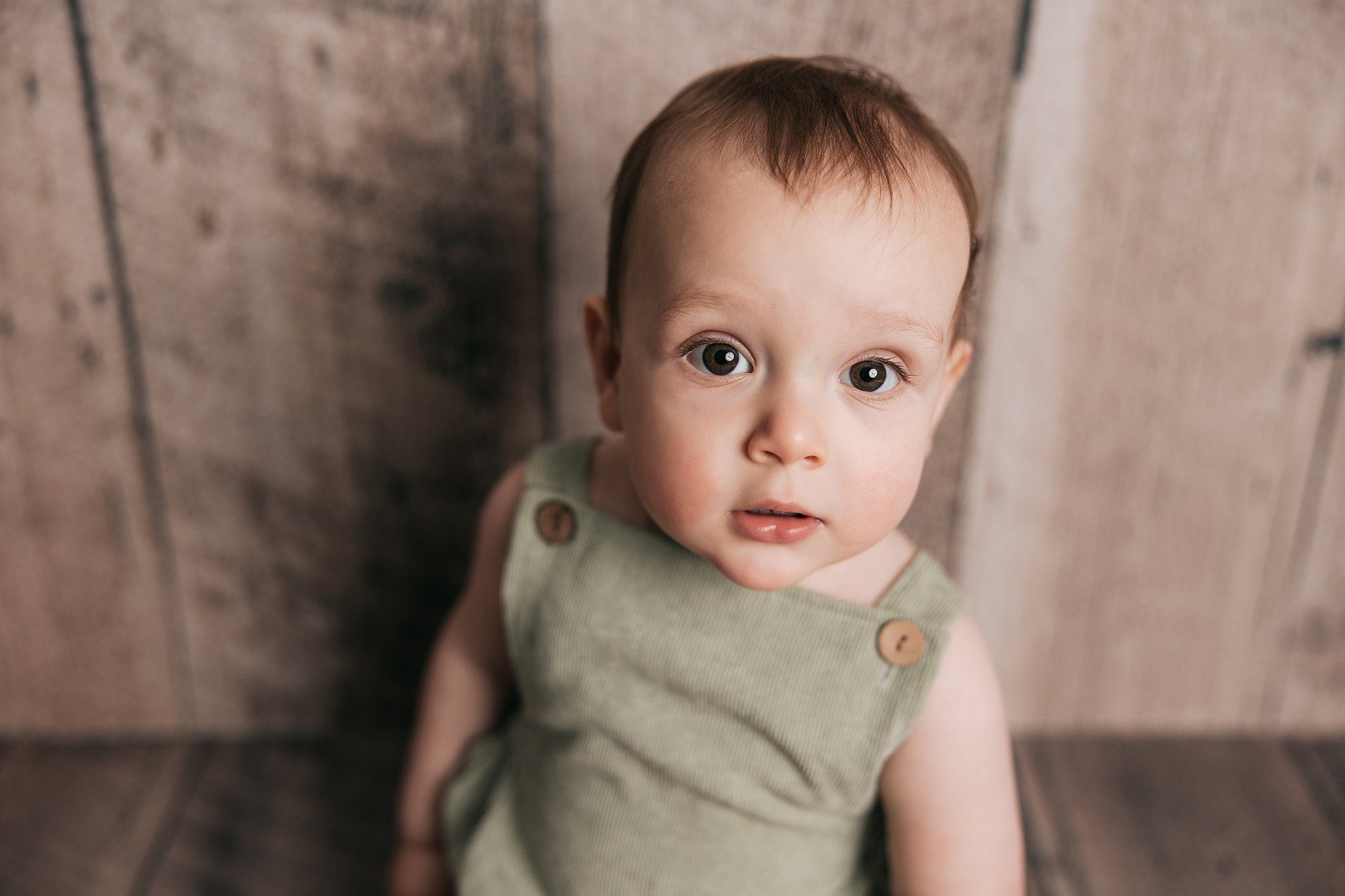A toddler boy in green overalls sits on the floor of a studio with eyes wide open