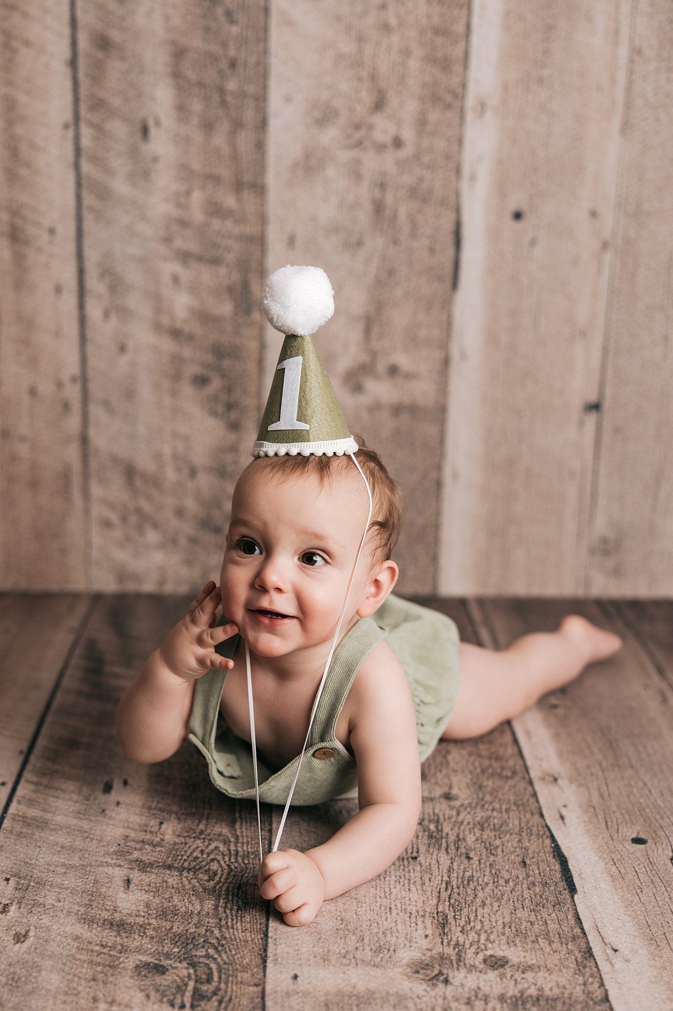 A toddler boy in green overalls lays on a floor smiling in a birthday hat before visiting a toy store raleigh nc