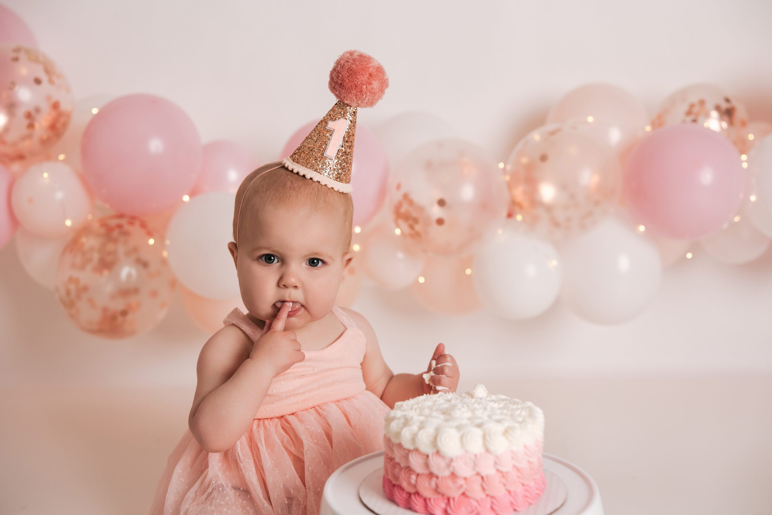 baby girl eating a cake for her first birthday with a party hat on, pink cake smash raleigh